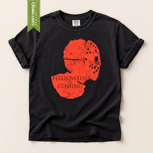 May include: A black short-sleeved t-shirt featuring a distressed red graphic of a hockey mask silhouette. The text "HALLOWEEN IS COMING" is printed in black within the red graphic. The shirt has a crew neckline and a Comfort Colors brand label.