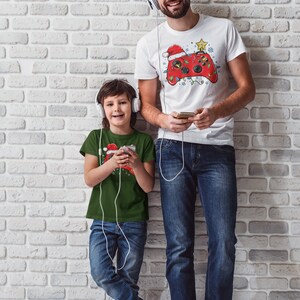 K&ouml;nnte beinhalten: Ein Vater und sein Sohn tragen passende Weihnachts-T-Shirts mit einem Videospiel-Controller-Design. Der Vater tr&auml;gt ein wei&szlig;es T-Shirt mit einem roten Controller und der Sohn tr&auml;gt ein gr&uuml;nes T-Shirt mit einem roten Controller. Beide tragen Kopfh&ouml;rer und Jeans.