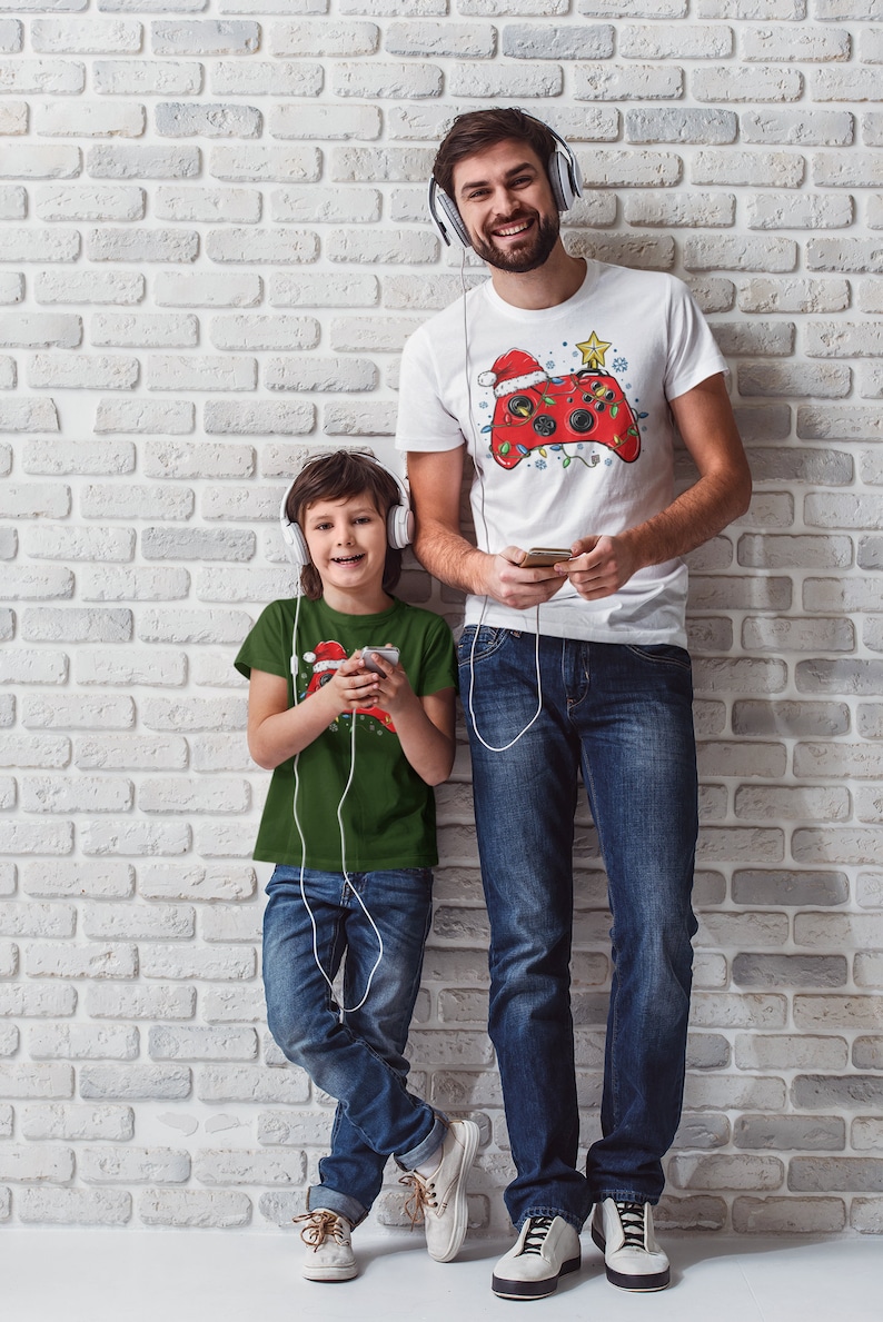 K&ouml;nnte beinhalten: Ein Vater und sein Sohn tragen passende Weihnachts-T-Shirts mit einem Videospiel-Controller-Design. Der Vater tr&auml;gt ein wei&szlig;es T-Shirt mit einem roten Controller und der Sohn tr&auml;gt ein gr&uuml;nes T-Shirt mit einem roten Controller. Beide tragen Kopfh&ouml;rer und Jeans.