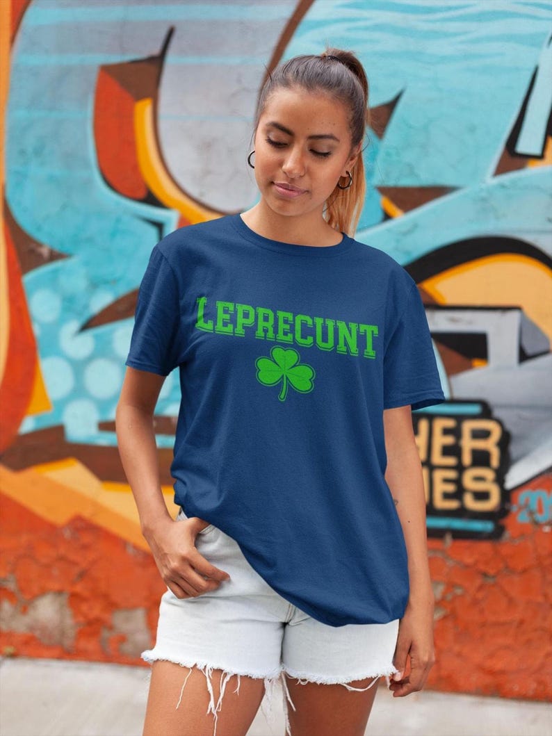 A young woman wearing a blue t-shirt with the word "Leprecht" printed on it, standing in front of a colorful graffiti wall.