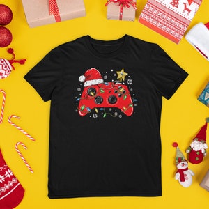 K&ouml;nnte beinhalten: Schwarzes T-Shirt mit einem roten Videospiel-Controller, der einen Weihnachtsmann-Hut tr&auml;gt und von Weihnachtslichtern umgeben ist. Der Controller befindet sich auf einem gelben Hintergrund mit Weihnachtsdekoration.