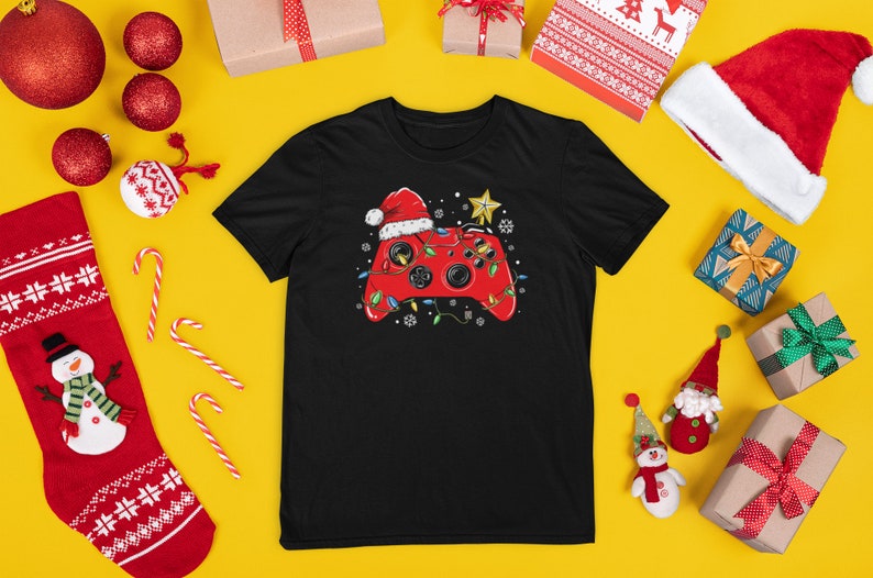 K&ouml;nnte beinhalten: Schwarzes T-Shirt mit einem roten Videospiel-Controller, der einen Weihnachtsmann-Hut tr&auml;gt und von Weihnachtslichtern umgeben ist. Der Controller befindet sich auf einem gelben Hintergrund mit Weihnachtsdekoration.
