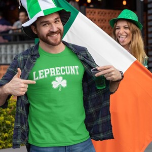 A man wearing a green shirt with the word " Leprechaun" and a large Irish flag is smiling and holding a drink, while a woman in a green hat is also smiling and holding an orange flag.