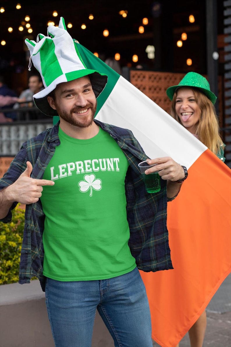 A man wearing a green shirt with the word " Leprechaun" and a large Irish flag is smiling and holding a drink, while a woman in a green hat is also smiling and holding an orange flag.