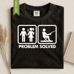 以下が含まれることがあります： 黒いTシャツで、2つの正方形のアイコンが白くプリントされています。最初のアイコンは誰かに話しかけている人物、2つ目のアイコンは釣りをする人物を描いています。アイコンの下には「PROBLEM SOLVED」という白い文字がプリントされています。