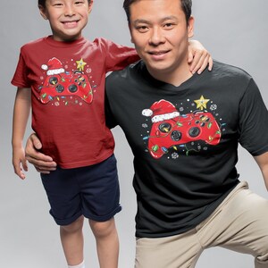 K&ouml;nnte beinhalten: Ein rot-schwarzes T-Shirt mit einem weihnachtlichen Videospiel-Controller-Design. Der Controller ist mit einem Weihnachtsmann-Hut, Lichtern und Schneeflocken verziert. Das Shirt ist in Erwachsenen- und Kindergr&ouml;&szlig;en erh&auml;ltlich.