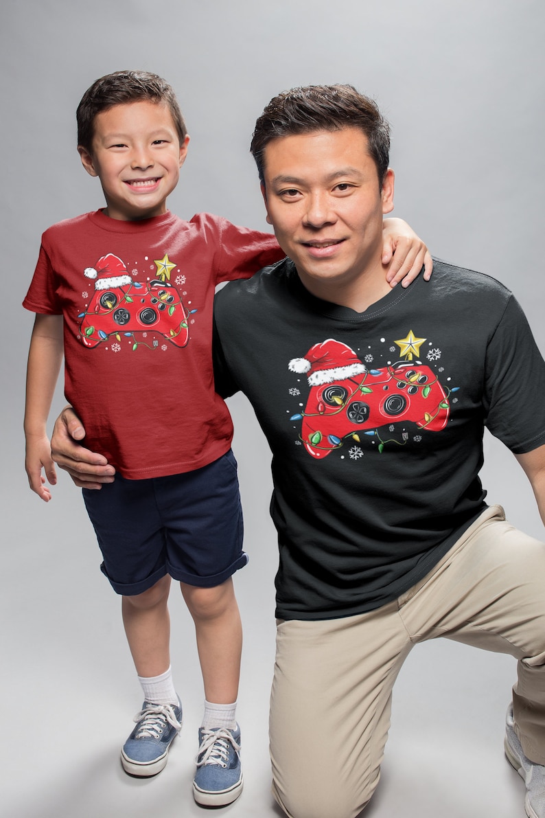 K&ouml;nnte beinhalten: Ein rot-schwarzes T-Shirt mit einem weihnachtlichen Videospiel-Controller-Design. Der Controller ist mit einem Weihnachtsmann-Hut, Lichtern und Schneeflocken verziert. Das Shirt ist in Erwachsenen- und Kindergr&ouml;&szlig;en erh&auml;ltlich.