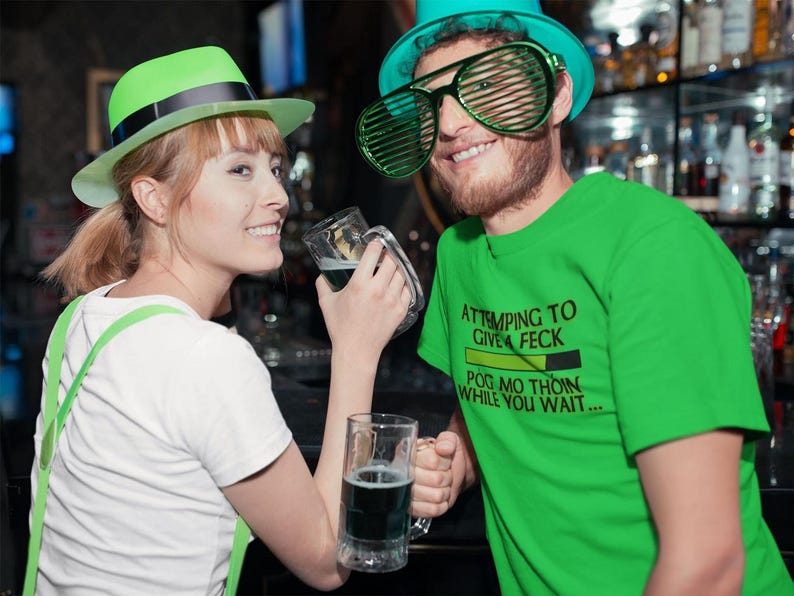 A woman in a white shirt and green hat is smiling and holding a glass of beer, while a man in a green t-shirt and large green glasses is also smiling and holding a glass of beer.