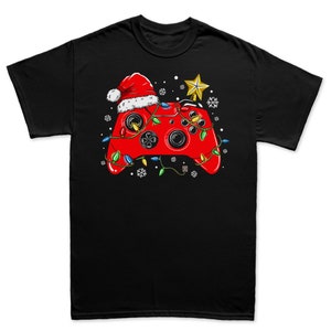 K&ouml;nnte beinhalten: Ein schwarzes T-Shirt mit einem roten Videospiel-Controller, der einen Weihnachtsmann-Hut und Weihnachtslichter tr&auml;gt. Der Controller ist von Schneeflocken umgeben.
