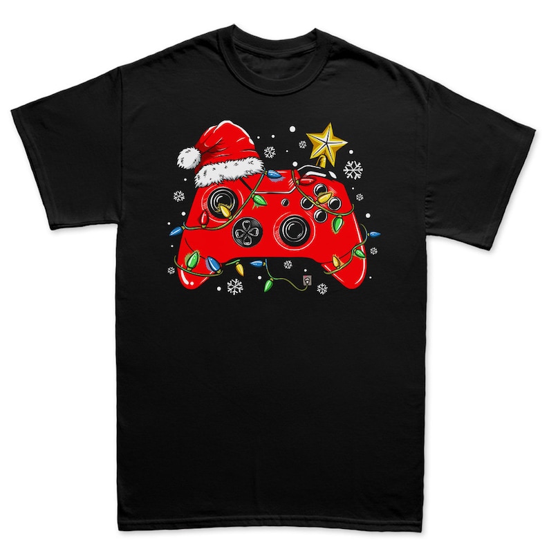 K&ouml;nnte beinhalten: Ein schwarzes T-Shirt mit einem roten Videospiel-Controller, der einen Weihnachtsmann-Hut und Weihnachtslichter tr&auml;gt. Der Controller ist von Schneeflocken umgeben.