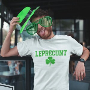 A man wearing a white t-shirt with the word "LEPRECIUT" and a shamrock symbol is posing with a green hat and large green glasses, making a playful and humorous expression.