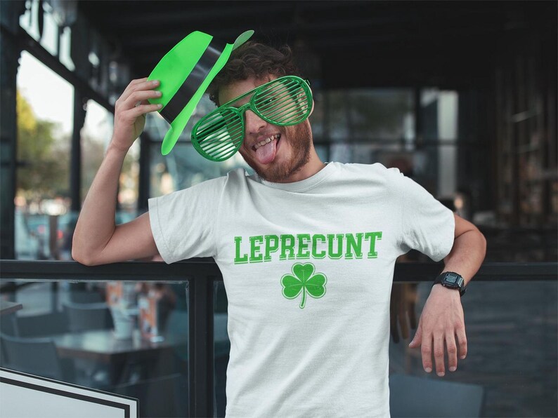 A man wearing a white t-shirt with the word "LEPRECIUT" and a shamrock symbol is posing with a green hat and large green glasses, making a playful and humorous expression.