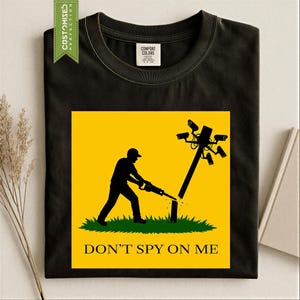 May include: A black crew neck t-shirt featuring a yellow square graphic. The graphic shows a silhouette of a person cutting down a utility pole topped with security cameras using a chainsaw. The text "DON'T SPY ON ME" is printed in black below.