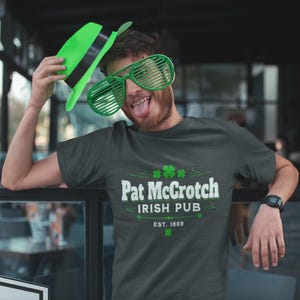 A man wearing a green hat and large sunglasses is posing for a photo in front of a sign for Pat Crotch Irish Pub.