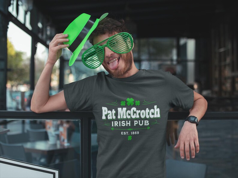A man wearing a green hat and large sunglasses is posing for a photo in front of a sign for Pat Crotch Irish Pub.