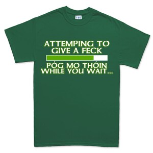 May include: A dark green t-shirt with white text that reads "ATTEMPTING TO GIVE A FECK" above a loading bar graphic, followed by "PÓG MO THÓIN WHILE YOU WAIT...". The shirt has a crew neck and a "TEE" tag.