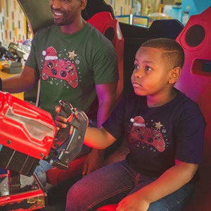 K&ouml;nnte beinhalten: Ein Vater und sein Sohn tragen passende gr&uuml;ne und schwarze T-Shirts mit einem weihnachtlichen Videospiel-Controller-Design. Die Shirts zeigen einen roten und gr&uuml;nen Controller mit einer Weihnachtsm&uuml;tze und Lichtern. Der Vater h&auml;lt einen roten Joystick und der Sohn schaut nach oben.