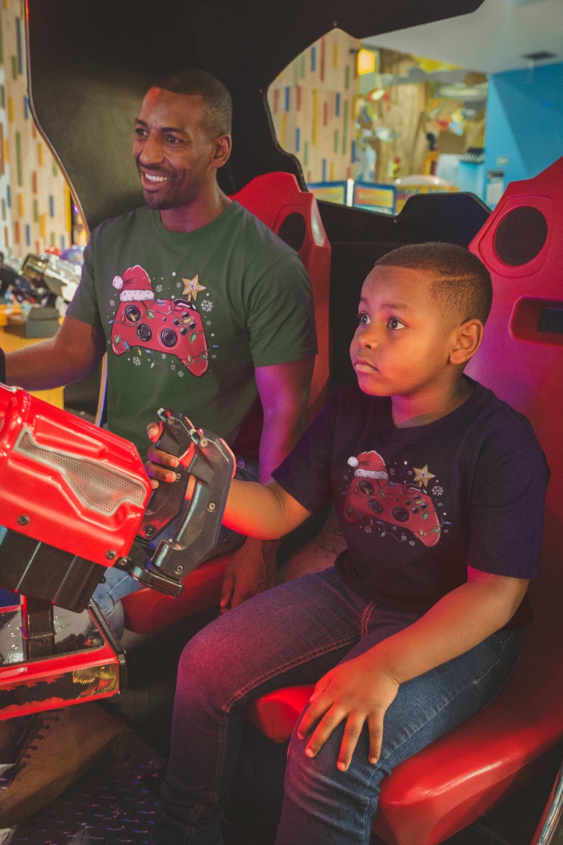 K&ouml;nnte beinhalten: Ein Vater und sein Sohn tragen passende gr&uuml;ne und schwarze T-Shirts mit einem weihnachtlichen Videospiel-Controller-Design. Die Shirts zeigen einen roten und gr&uuml;nen Controller mit einer Weihnachtsm&uuml;tze und Lichtern. Der Vater h&auml;lt einen roten Joystick und der Sohn schaut nach oben.