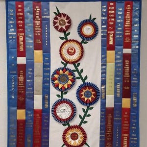 May include: A wall hanging featuring a white background with a floral design of rosettes in red, blue, and gold. Vertical blue and red ribbons with text are on either side, with the word "Champion" prominently displayed.