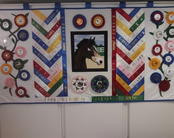May include: A white quilt featuring a horse portrait in the centre. The quilt is decorated with blue, red, yellow and green ribbons, and a variety of rosettes. The quilt has the words "FOURTH" and "SIXTH" written on it.