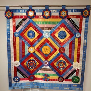 May include: A colorful ribbon quilt featuring rosettes and ribbons in red, blue, yellow, and white. The quilt is adorned with ribbons that say "Champion", "First Place", and other awards. The quilt is a decorative piece, likely for a competition.