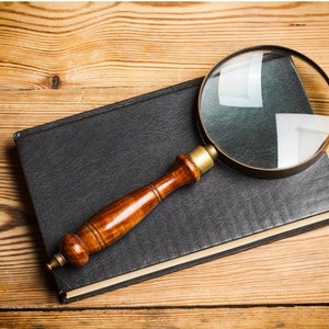 Op de afbeelding: Een vergrootglas met een houten handvat ligt op een zwart lederen gebonden boek. Het boek en het vergrootglas liggen op een houten oppervlak.