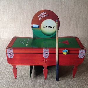 May include: A green felt snooker table with a red wooden frame and white felt pockets. The table has a cue stick and a white snooker ball with the name "GARRY" on it. A sign above the table says "Happy Birthday!"