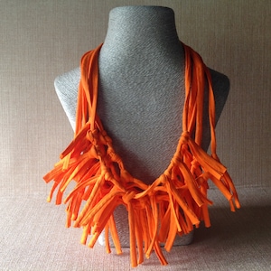 May include: Bright orange fabric necklace displayed on a mannequin. The necklace is made of knotted strips of fabric, creating a fringed, statement piece. The vibrant color and unique design make it a bold accessory.