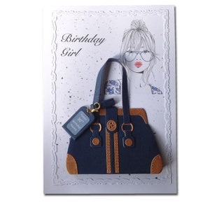 Op de afbeelding: Een blauwe en bruine handtas met een gouden ketting en een label met de tekst "Happy 30th Birthday Hayley". De handtas is op een witte kaart met een tekening van een vrouw met een knot en een bril.