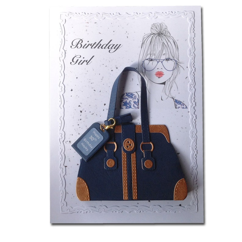 Op de afbeelding: Een blauwe en bruine handtas met een gouden ketting en een label met de tekst "Happy 30th Birthday Hayley". De handtas is op een witte kaart met een tekening van een vrouw met een knot en een bril.