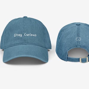 May include: A denim blue baseball cap with the phrase "Stay Curious" embroidered in white on the front. The back of the cap features a small smiley face. The cap has an adjustable strap.