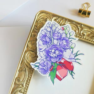 May include: A sticker featuring a bouquet of purple and white flowers with a large red gemstone in the center. The flowers are drawn in a delicate style with a white background.