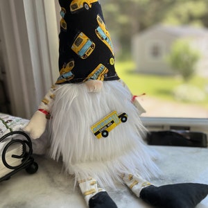 May include: A white plush gnome with a black hat featuring a yellow school bus pattern. The gnome is wearing white pants with a yellow school bus pattern and black shoes. The gnome has a white beard and a red nose. The gnome is sitting on a white surface.