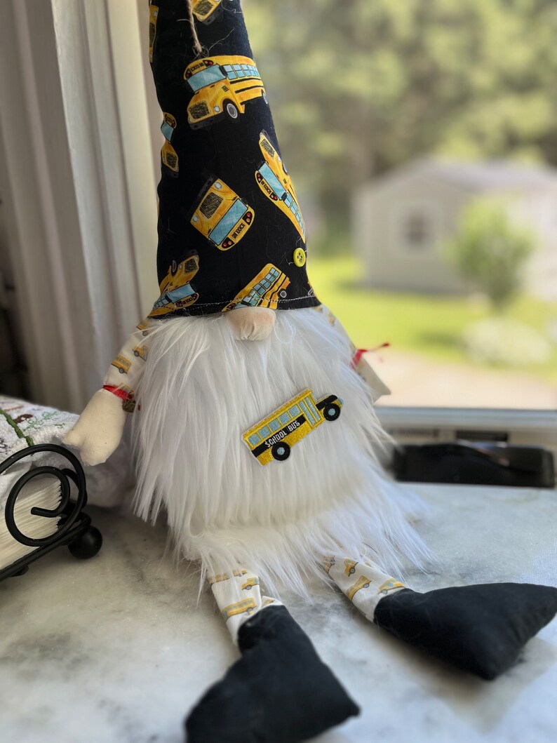 May include: A white plush gnome with a black hat featuring a yellow school bus pattern. The gnome is wearing white pants with a yellow school bus pattern and black shoes. The gnome has a white beard and a red nose. The gnome is sitting on a white surface.