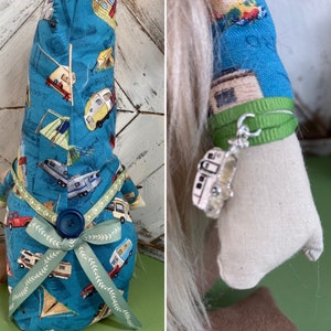 May include: A blue gnome with a white beard and a green ribbon around its waist. The gnome is wearing a blue and white fabric with a pattern of campers and tents. The gnome is holding a silver charm of a camper.