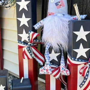 May include: Four wooden blocks painted in red, white, and blue with white stars. The blocks are decorated with red, white, and blue ribbon. A gnome wearing a grey hat with red, white, and blue stars and a white beard is standing in front of the blocks.