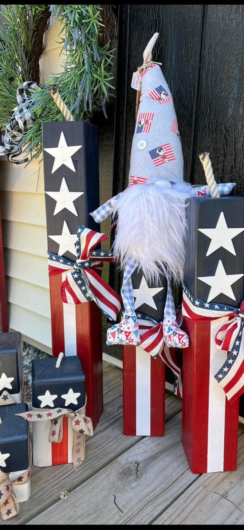 May include: Four wooden blocks painted in red, white, and blue with white stars. The blocks are decorated with red, white, and blue ribbon. A gnome wearing a grey hat with red, white, and blue stars and a white beard is standing in front of the blocks.