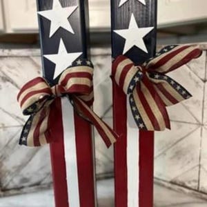 May include: Two wooden blocks painted in red, white, and blue with stars and stripes. Each block has a red, white, and blue ribbon tied around it.