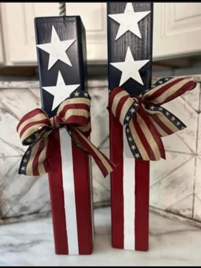 May include: Two wooden blocks painted in red, white, and blue with stars and stripes. Each block has a red, white, and blue ribbon tied around it.