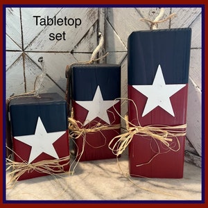 May include: Tabletop set of three wooden blocks decorated with a red, white, and blue American flag design. Each block is tied with a natural twine bow.
