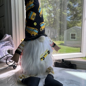 May include: A white gnome with a black and yellow hat featuring a school bus pattern. The gnome is wearing black pants with a yellow school bus patch and has a white beard.