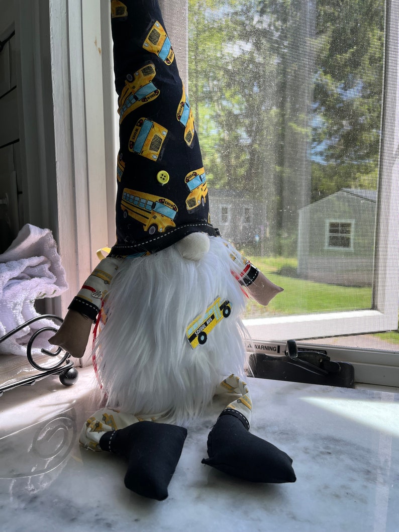 May include: A white gnome with a black and yellow hat featuring a school bus pattern. The gnome is wearing black pants with a yellow school bus patch and has a white beard.