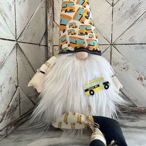 May include: A white gnome with a long, pointed hat covered in a yellow and blue school bus pattern. The gnome has a white beard and is wearing a black and white school bus patch on its chest. The gnome is sitting on a white surface.