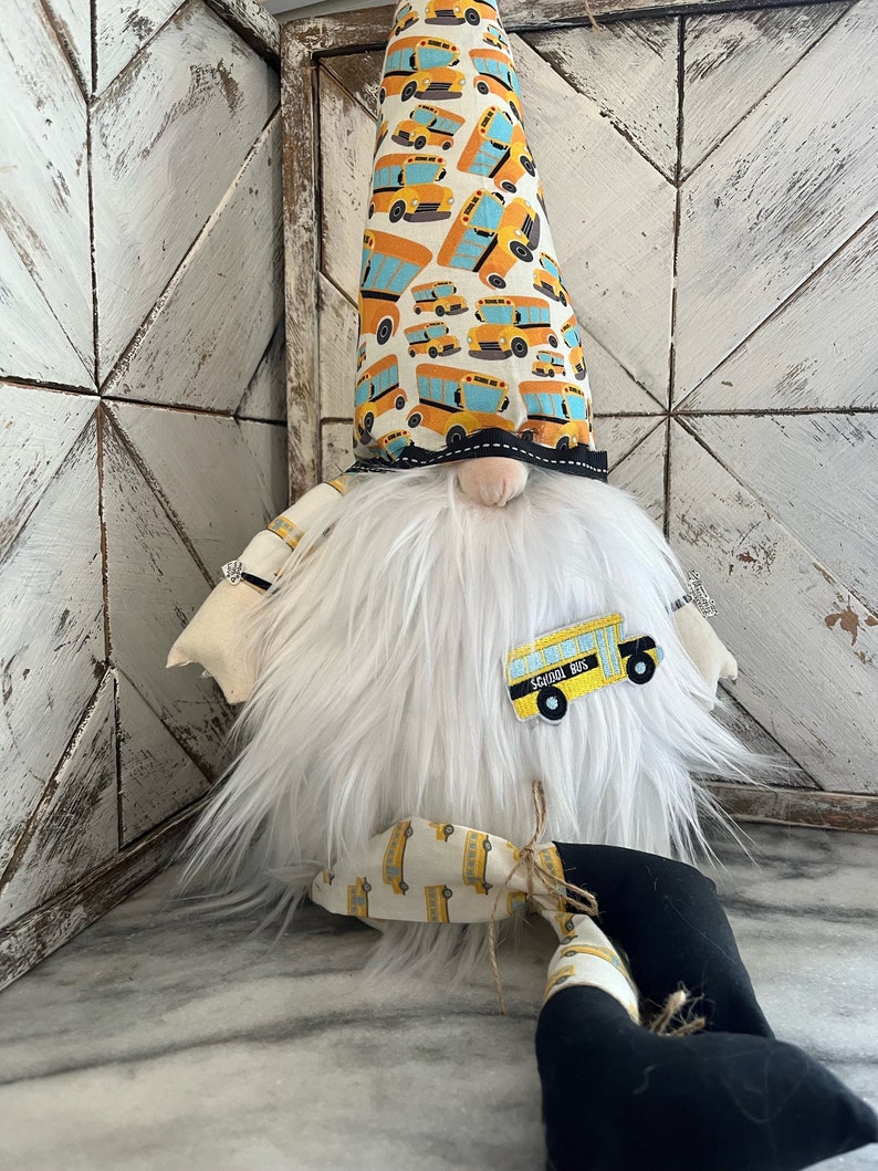May include: A white gnome with a long, pointed hat covered in a yellow and blue school bus pattern. The gnome has a white beard and is wearing a black and white school bus patch on its chest. The gnome is sitting on a white surface.