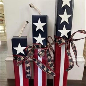 May include: Three wooden blocks painted in red, white, and blue with white stars. The blocks are decorated with a red, white, and blue ribbon. The blocks are arranged in a row from largest to smallest.