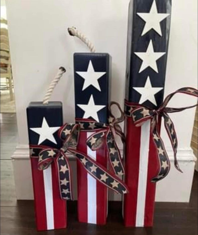 May include: Three wooden blocks painted in red, white, and blue with white stars. The blocks are decorated with a red, white, and blue ribbon. The blocks are arranged in a row from largest to smallest.