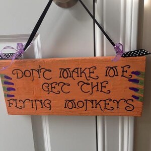 May include: Orange wooden sign with black lettering saying "DON'T MAKE ME GET THE FLYING MONKEYS." The sign has purple and green accents and black ribbon. It is hanging on a white door.