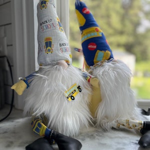 May include: Two gnomes wearing white fur and hats with a school bus theme. The hats have the text "Back to School" on them. The gnomes are sitting on a white surface.