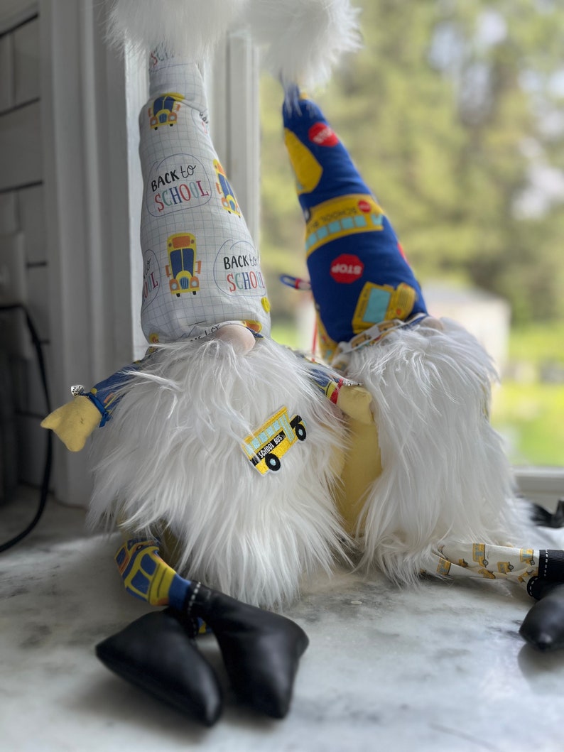 May include: Two gnomes wearing white fur and hats with a school bus theme. The hats have the text "Back to School" on them. The gnomes are sitting on a white surface.