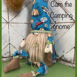 May include: A blue gnome with a white beard and a hat with a camping theme. The hat has a pattern of tents, campers, and cars. The gnome is sitting on a green surface. The text "Cam the Camping Gnome" is written in white on a wooden background.